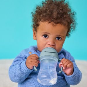 Dziecko z kręconymi włosami w niebieskim sweterku siedzi na turkusowym tle, trzymając i pijąc z bidonu ze słomką 240 ml Ocean, patrząc bezpośrednio w kamerę.