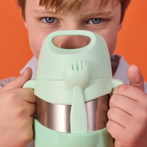 Dziecko trzyma b.box Termos na jedzenie, 335 ml, Spearmint, z pasującym widelcem przymocowanym z przodu. Pojemnik częściowo zasłania twarz dziecka i wyróżnia się na pomarańczowym tle kontrastującym z jego głębokim odcieniem.