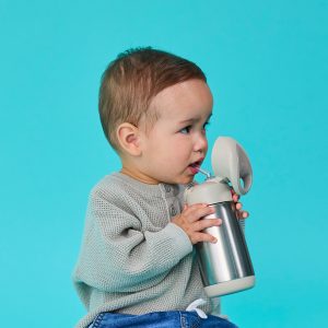 Małe dziecko z krótkimi brązowymi włosami, ubrane w jasnoszary sweter i niebieskie dżinsy, pije z butelki Bidon Termiczny o pojemności 350 ml w kolorze Latte, z pokrywką typu flip top, na jednolitym turkusowym tle. Dziecko patrzy w bok, trzymając butelkę.