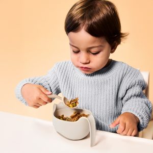 Małe dziecko w jasnoszarym sweterku siedzi przy białym stole, uważnie jedząc z trzymanej miski za pomocą b.box Pierwsze sztućce dla niemowląt, Latte. Tło ma ciepły brzoskwiniowy odcień.