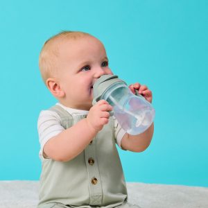 Uśmiechnięte dziecko z krótkimi blond włosami, w jasnozielonym kombinezonie i białej koszuli, siedzi na miękkiej powierzchni na jasnoniebieskim tle, trzymając i pijąc z bidon ze słomką 240 ml Sage z zieloną pokrywką.