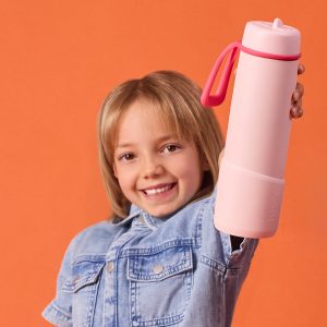 Dziewczynka uśmiecha się i prezentuje butelkę b.box 690 ml z nakładką w kolorze Pink Paradise, stojąc na pomarańczowym tle.